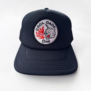 May include: A navy blue trucker hat with a curved peak and mesh back. The front displays a circular patch with the text "COOL DADS PUNS CLUB" around a cartoon unicorn-tiger design in red, white, and black.
