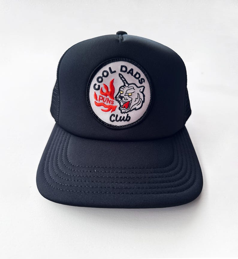 May include: A navy blue trucker hat with a curved peak and mesh back. The front displays a circular patch with the text "COOL DADS PUNS CLUB" around a cartoon unicorn-tiger design in red, white, and black.