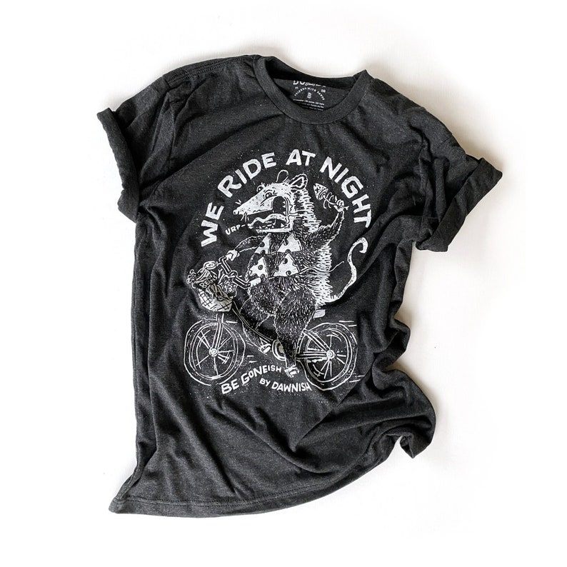 May include: A dark grey t-shirt with a white graphic of a possum riding a bicycle. The graphic includes the text "We Ride At Night" and "Be Gone-ish By Dawn-ish".