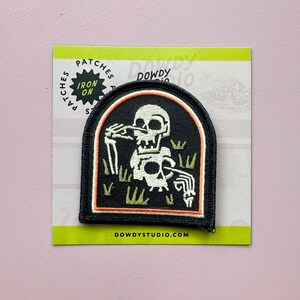 May include: Black and white iron-on patch featuring a skeleton in a graveyard scene. The patch has a black border with an orange inner border. The text "IRON ON" is on the top left corner. The website address DOWDYSTUDIO.COM is at the bottom.