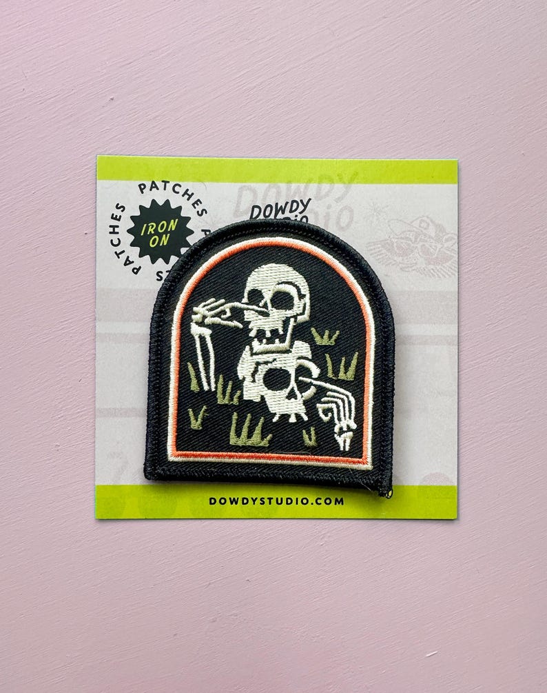 May include: Black and white iron-on patch featuring a skeleton in a graveyard scene. The patch has a black border with an orange inner border. The text "IRON ON" is on the top left corner. The website address DOWDYSTUDIO.COM is at the bottom.