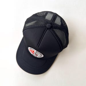 May include: Black trucker cap with a mesh back and a curved peak. The cap has a patch with a tiger graphic and the text "Real Bad Club". A button is on top.