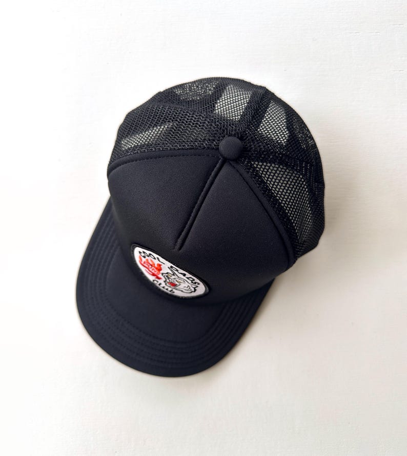 May include: Black trucker cap with a mesh back and a curved peak. The cap has a patch with a tiger graphic and the text "Real Bad Club". A button is on top.