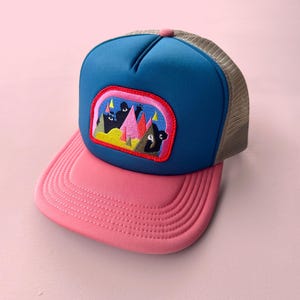 Puede incluir: Gorra de camionero con panel frontal azul, visera rosa y parte trasera de malla beige. La gorra presenta un parche bordado con una colorida escena de montaña. El botón superior es rosa.