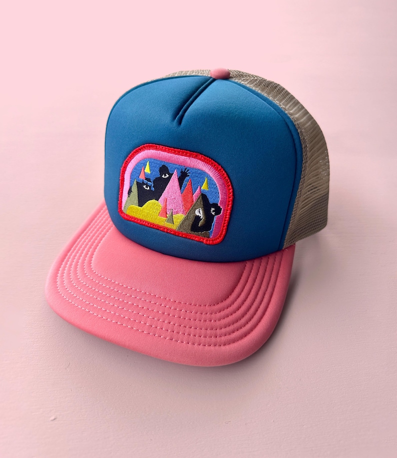 May include: A trucker hat with a blue front panel, pink brim, and beige mesh back. The hat features an embroidered patch with a colourful mountain scene. The top button is pink.
