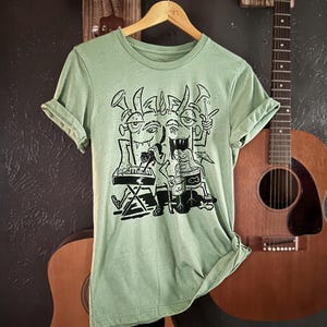 Op de afbeelding: Een lichtgroen T-shirt met een zwart-witte grafische print van drie cartoonmonsters die muziek maken. De monsters dragen hoeden en hebben hoorns. E&eacute;n monster speelt keyboard, een ander speelt drums en de derde speelt trompet.
