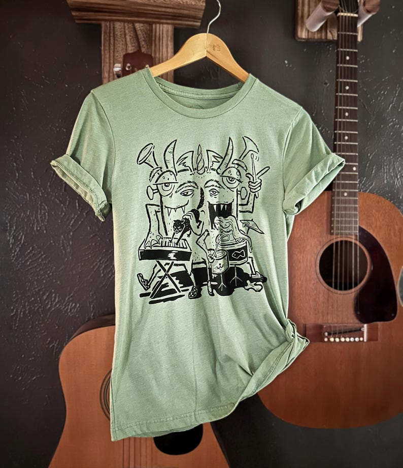 Op de afbeelding: Een lichtgroen T-shirt met een zwart-witte grafische print van drie cartoonmonsters die muziek maken. De monsters dragen hoeden en hebben hoorns. E&eacute;n monster speelt keyboard, een ander speelt drums en de derde speelt trompet.