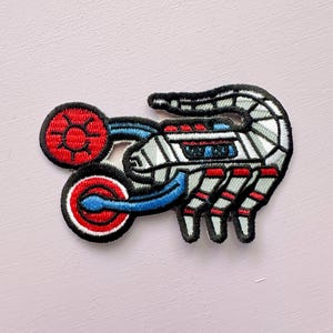 May include: Embroidered patch featuring a stylized, mechanical design. The patch has a black outline with red, white, and blue accents. The design includes a circular element and a segmented, insect-like body.