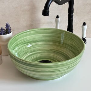 May include: A green and white swirl-patterned ceramic sink bowl with a black tap and white and black handles. A small white vase with purple flowers and a beige candle are to the left of the sink.