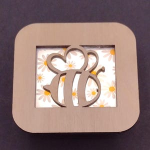 May include: Two wooden square frames with a cutout of a bee with yellow and black stripes on a white background with a daisy pattern.