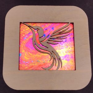 May include: Two wooden frames with a hummingbird cutout. The frames are made of light wood and have a square shape. The background of the frames is a vibrant pink and blue abstract design.