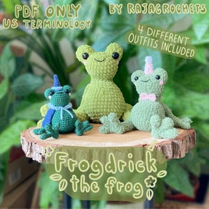 May include: Three green crocheted frog toys are displayed on a wooden slice. One frog has a blue cape and hat, another a pink bow tie, and the third is plain. The image includes the text "Frogdrick the frog" and "4 different outfits included."