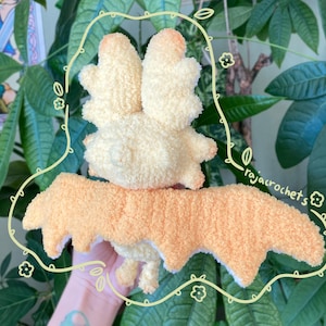 May include: A handmade, plush bat toy with a cream-colored body and peach-colored accents. The bat has large, fluffy wings and a textured, soft appearance. The toy is likely crocheted or knitted, and is designed to be a cuddly toy.