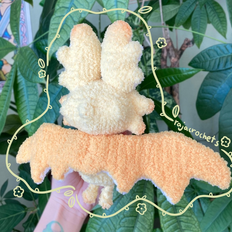 May include: A handmade, plush bat toy with a cream-colored body and peach-colored accents. The bat has large, fluffy wings and a textured, soft appearance. The toy is likely crocheted or knitted, and is designed to be a cuddly toy.
