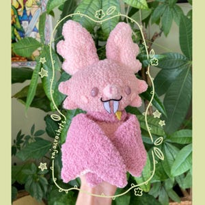 May include: A handmade pink plush bat with large, textured wings and a small, smiling face. The bat has a black mustache, white fangs, and a yellow tongue. The plushie is surrounded by a decorative border with the text "rajacrochets".