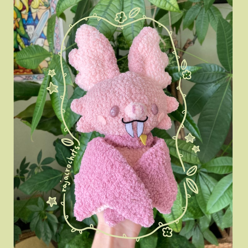 May include: A handmade pink plush bat with large, textured wings and a small, smiling face. The bat has a black mustache, white fangs, and a yellow tongue. The plushie is surrounded by a decorative border with the text "rajacrochets".