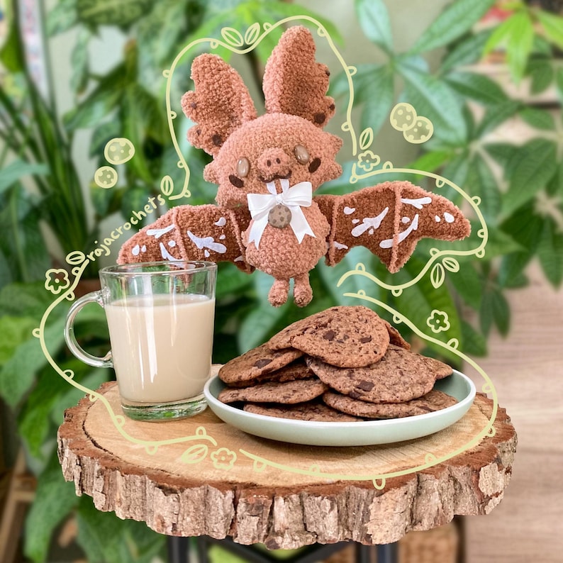 May include: A crocheted brown bat plush toy with a white bow and a small cookie. The bat is above a plate of chocolate chip cookies and a glass mug of a light-colored beverage. The image is set on a wooden slice.