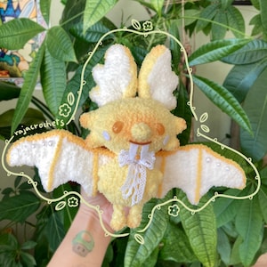 May include: A handmade crochet moth plushie in shades of yellow and white. The plushie has large, fluffy wings with pearl accents and a delicate lace bow. The text "rajacrochets" is visible. The moth is a whimsical, soft toy.