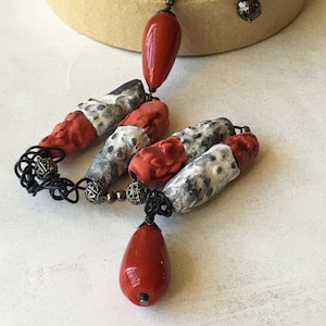 May include: A necklace with a long chain and multiple red and white ceramic beads. The beads are teardrop shaped and have a speckled pattern. The necklace is finished with a small metal bead at the end.