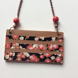 May include: A brown wooden sign with a black background and red and white floral pattern. The sign has three horizontal stripes of the floral pattern and a circle and rectangle in the middle. The sign is hanging from a copper chain with red beads.