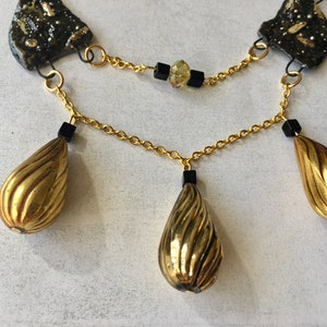 Könnte beinhalten: Eine goldene Halskette mit drei tropfenförmigen Anhängern, schwarzen Perlen und einer goldenen Kette. Die Anhänger haben eine strukturierte, gerippte Oberfläche.