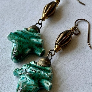 May include: Handmade earrings with turquoise ceramic pendants. The pendants have a textured surface and are suspended from brass-coloured metal beads and ear wires. The ceramic elements have an organic shape, adding to their unique design.
