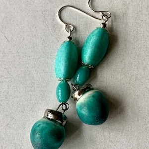 May include: A pair of drop earrings featuring turquoise-coloured beads in varying shapes, including elongated oval and rounded forms. The beads are accented with silver-toned metal caps and connected by silver-toned wire hooks for hanging.