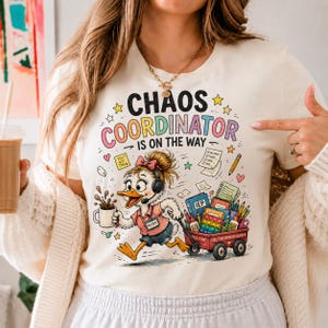 以下が含まれることがあります： ヘッドセットを装着しコーヒーマグを持った漫画風のアヒルのグラフィックが描かれたクリーム色のTシャツ。アヒルの上には「CHAOS COORDINATOR IS ON THE WAY」という文字があり、アヒルは学用品でいっぱいの赤いワゴンを引いています。