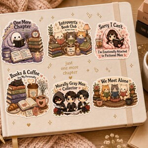 K&ouml;nnte beinhalten: Ein beiges Notizbuch mit Aufklebern mit Illustrationen zum Thema B&uuml;cher. Eine Kerze, ein Stift und eine Kaffeetasse sind ebenfalls zu sehen. Die Aufkleber tragen Texte wie "One More Chapter" und "Books & Coffee Are My Personality."