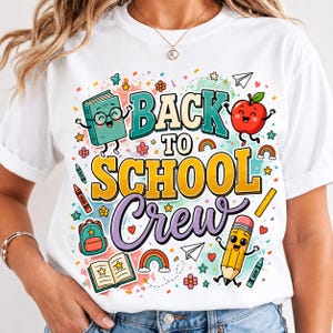 Può includere: Una t-shirt bianca con una grafica colorata e la scritta "BACK TO SCHOOL Crew". Il design include illustrazioni a cartoni animati di un libro sorridente, una mela rossa, una matita, uno zaino, un libro aperto, arcobaleni, pastelli e aeroplanini di carta.