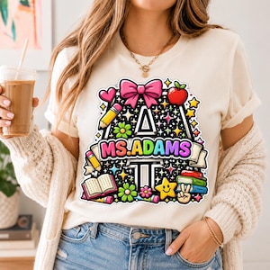 May include: Cream-colored t-shirt featuring a colorful graphic design with the text "MS.ADAMS". The design includes a large letter "A" filled with stars, flowers, and school-themed elements like pencils, books, and an apple.