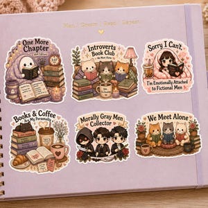 K&ouml;nnte beinhalten: Eine Sammlung farbenfroher Aufkleber auf einem Spiralnotizbuch. Die Aufkleber zeigen niedliche Illustrationen von Katzen, B&uuml;chern, Kaffee und S&auml;tzen wie "Introverts Book Club" und "Books & Coffee". Eine Kerze und ein Buch sind ebenfalls zu sehen.