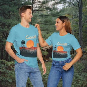 Puede incluir: Dos personas con camisetas azul claro con un gráfico de una puesta de sol y una torre con el texto "Catskills" en cada camiseta. Las personas están de pie en un entorno boscoso.