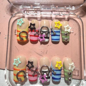 May include: A set of ten press-on nails arranged in a clear plastic case. Each nail features a nude base with horizontal stripes in yellow, pink, black, blue, and green. The nails are decorated with small colorful star charms and silver-toned metal hoop piercings.