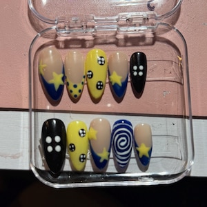May include: A set of ten artificial nails in a clear plastic case. The nails feature various designs, including yellow stars, blue crescent moons, white polka dots, and a spiral design. The nails are in a variety of colors, including yellow, blue, black, and nude.