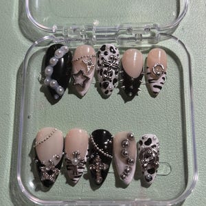 May include: A set of ten almond-shaped press-on nails in a clear plastic case. The nails feature a black and white color palette with designs including cow print, French tips, silver cross charms, star-shaped studs, pearl accents, and metallic beads.