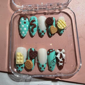 May include: A collection of ten artificial nails presented in a clear case. The nails are decorated with a variety of designs, including teal with white polka dots, chocolate swirls, and miniature food-themed decorations such as biscuits and waffles. The nails are oval-shaped.