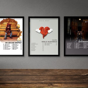May include: Three framed posters of Kanye West albums. The first poster is for "The College Dropout" and features a cartoon bear wearing a graduation cap. The second poster is for "808s & Heartbreak" and features a broken heart with hands holding it. The third poster is for "Late Registration" and features a dark image of a doorway.