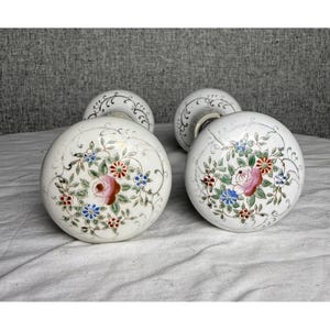 May include: Four white porcelain door knobs with floral designs. Each knob features a central pink rose surrounded by blue, red, and green flowers and leaves. The knobs have a decorative, curved base with gold accents.