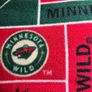 May include: A red, green, and white patterned fleece blanket. The blanket features the Minnesota Wild logo and the word "WILD" in large letters. The word "MINNESOTA" is also visible.