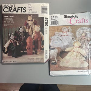 May include: Two craft pattern envelopes. The McCall's pattern features cat dolls with clothing, and the text "The Cat's Meow". The Simplicity pattern shows doll designs. Both patterns are for crafts.