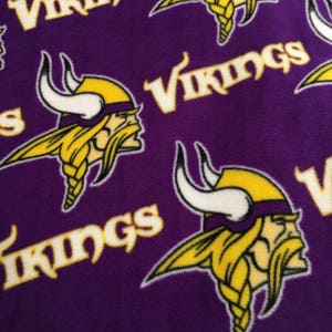 May include: Purple fabric with a repeating pattern of the word "Vikings" in gold and a Viking head logo. The Viking head features a yellow helmet, blonde braided beard, and white horns.