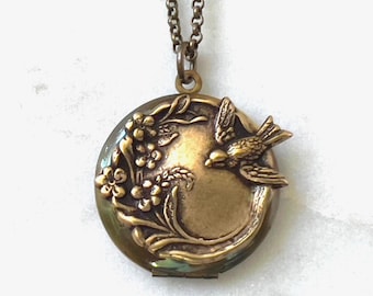 May include: A round brass locket pendant suspended from a matching metal chain. The front of the locket features a raised relief design depicting a small bird in flight alongside a cluster of flowers and foliage, all finished in a warm bronze tone.