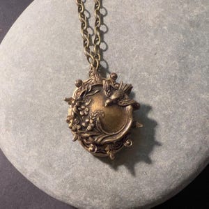 May include: A round bronze-toned metal locket pendant suspended from a matching link chain. The locket features a raised relief design depicting a small bird perched among detailed floral and leafy branches on its front surface.