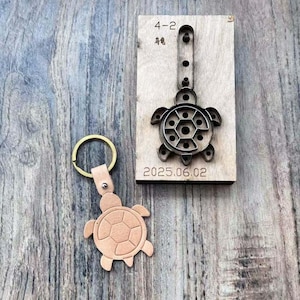 May include: A light brown leather turtle keychain next to a metal turtle-shaped die-cut tool. The die-cut tool is mounted on a wooden block with the date "2025.06.02" and the numbers "4-2" printed on it.