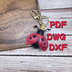 Puede incluir: Un llavero de mariquita con alas rojas y lunares negros, un cuerpo negro y herrajes dorados. El llavero está sobre una superficie de madera. El texto rojo dice "PDF", "DWG" y "DXF".