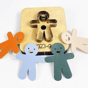 May include: A set of four leather gingerbread people cutouts in orange, blue, green, and beige. The cutouts are made using a metal die cutter with the number "023-9" visible on the wooden base.