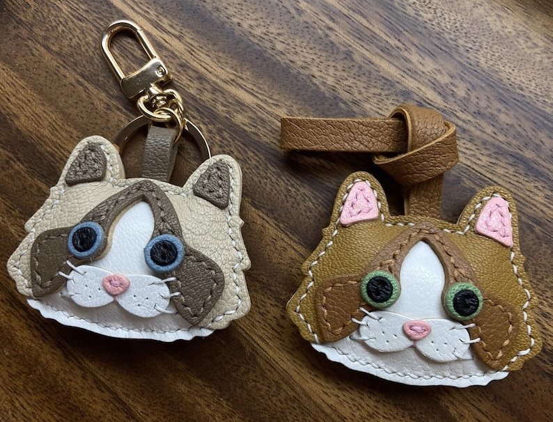 May include: Two handmade leather cat keychains. One is a brown tabby cat with green eyes and pink ears. The other is a white and brown cat with blue eyes. Both have stitched details and are attached to key rings.