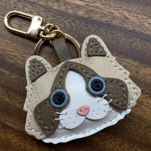 May include: A brown and white leather cat keychain with blue eyes and a pink nose. The keychain is attached to a gold metal ring.