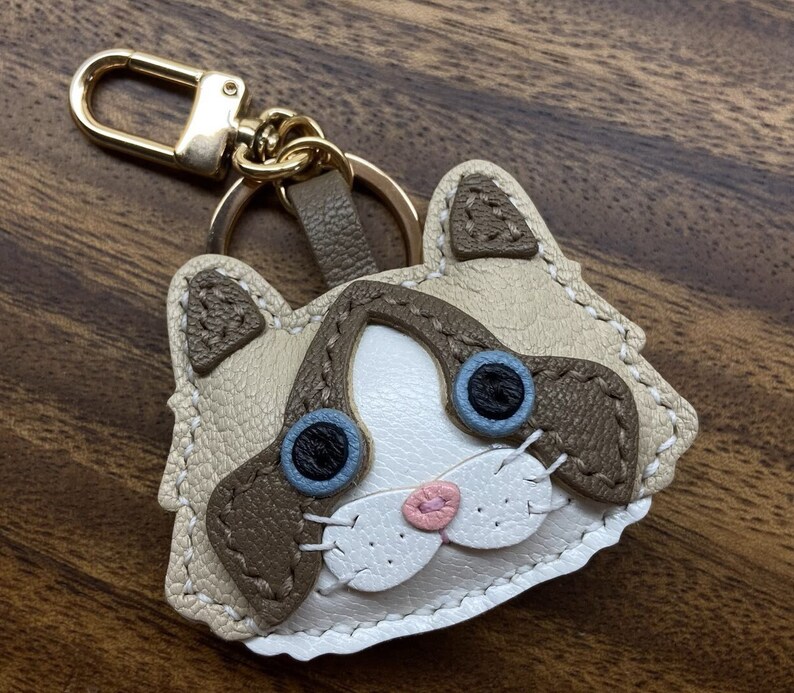 May include: A brown and white leather cat keychain with blue eyes and a pink nose. The keychain is attached to a gold metal ring.